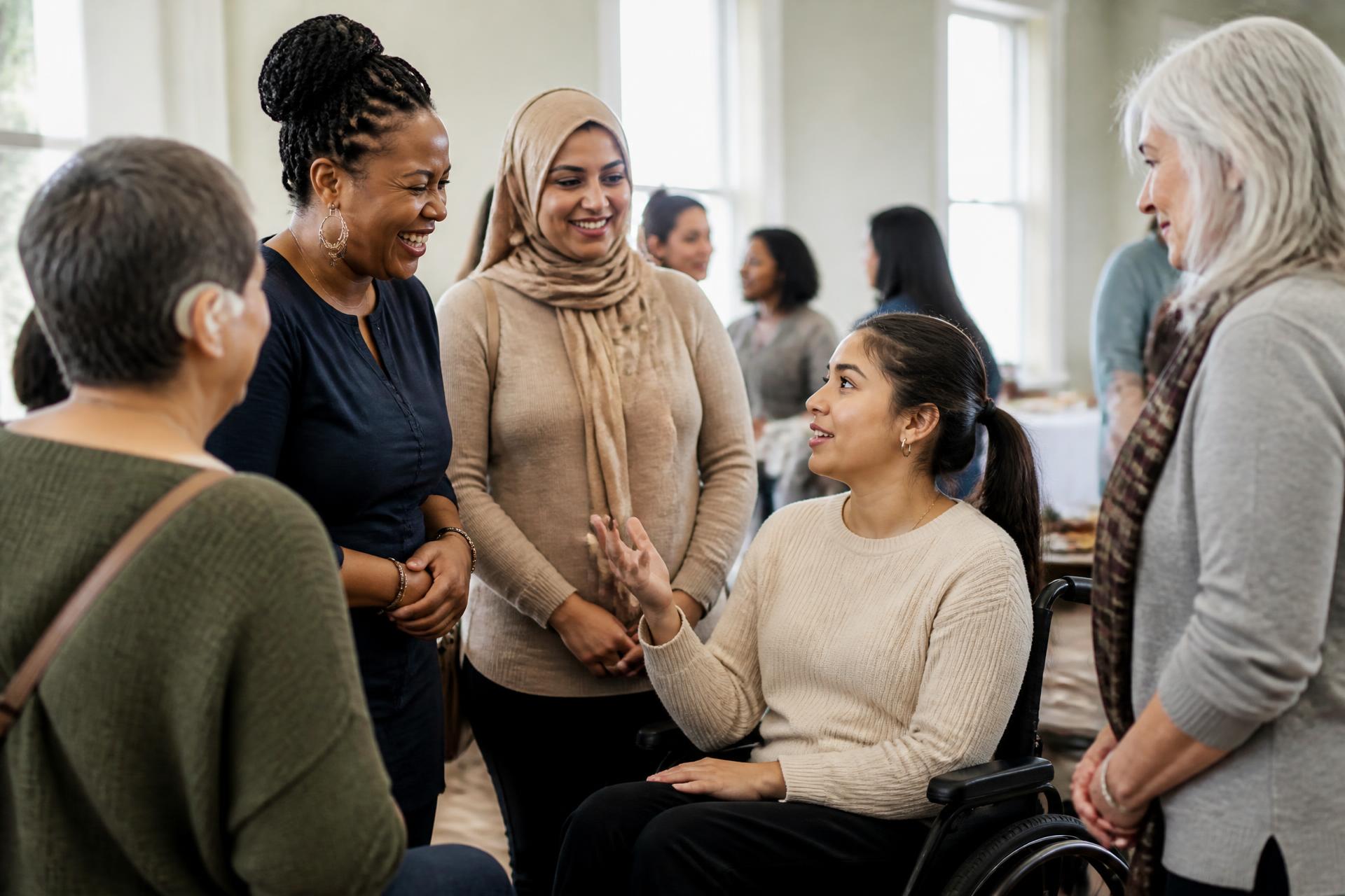 Women of different ages, ethnicities and abilities in conversation at a With Women Academy community event, including a wheelchair user speaking as an equal part of the group